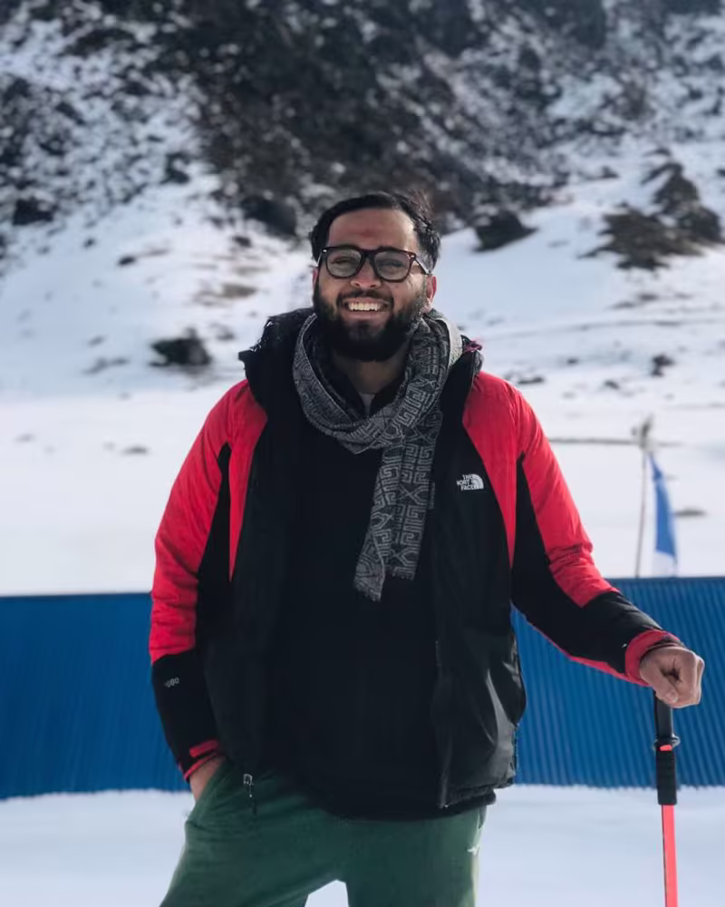 Pratik Poudel in a red trekking jacket and scarf, smiling with a trekking pole on a snowy mountainside