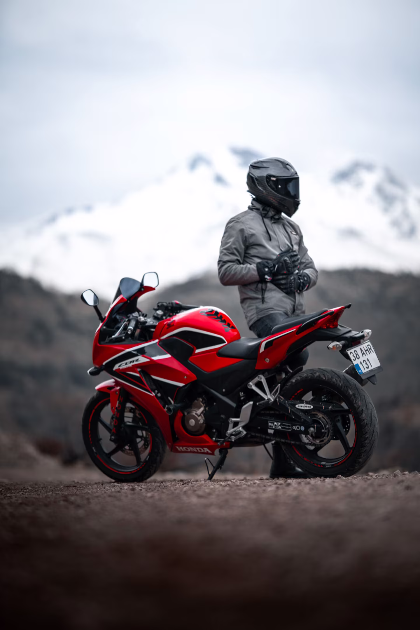 A rider standing beside their motorcycle below snow-capped mountains