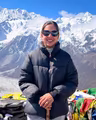 Dr. Barsha Dahal in a puffer jacket and sunglasses, smiling at a high mountain pass with snow peaks and prayer flags behind her
