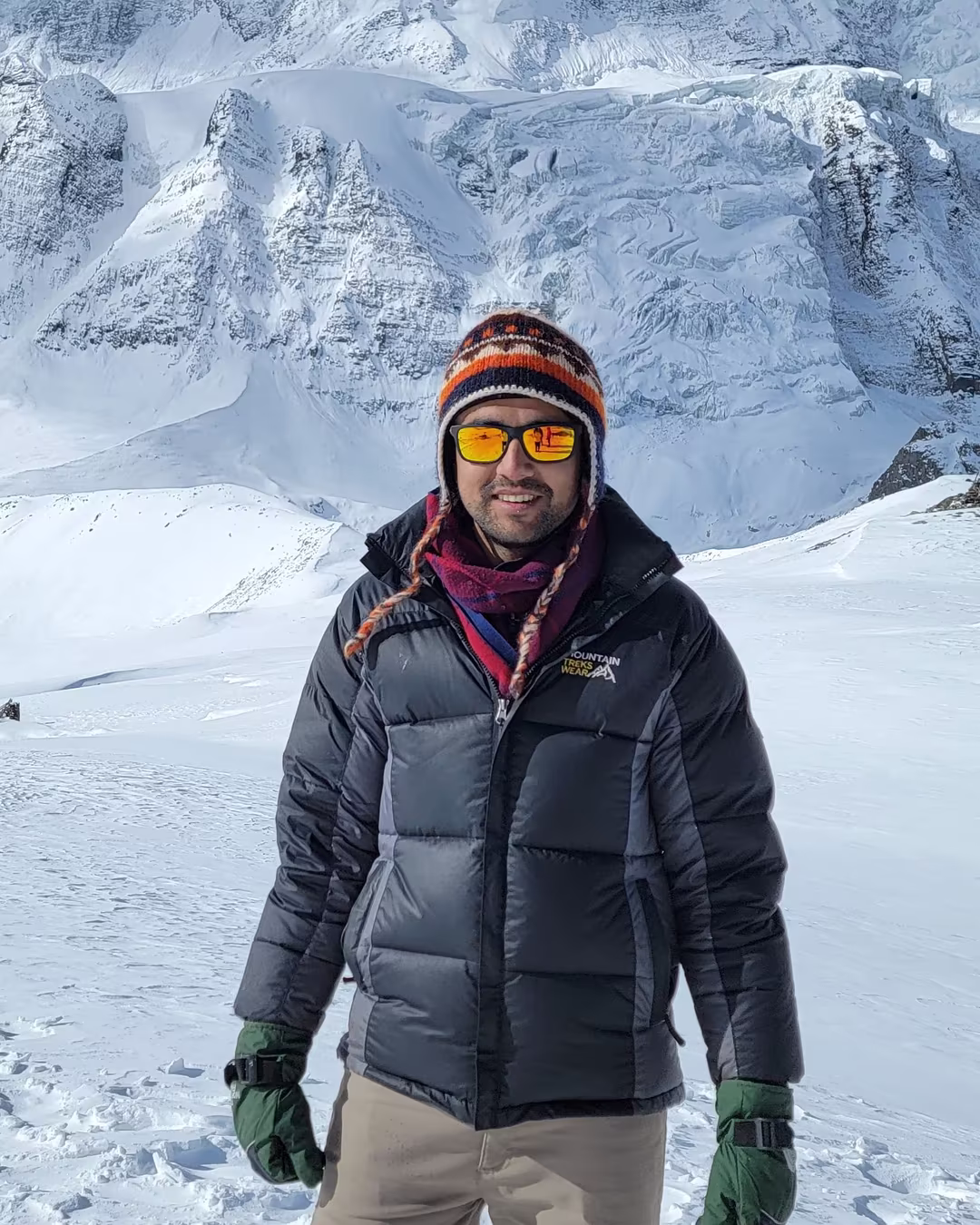 Sarun Luitel in a down jacket and ski goggles, smiling on a snow field below a steep Himalayan wall