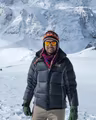 Sarun Luitel in a down jacket and ski goggles, smiling on a snow field below a steep Himalayan wall