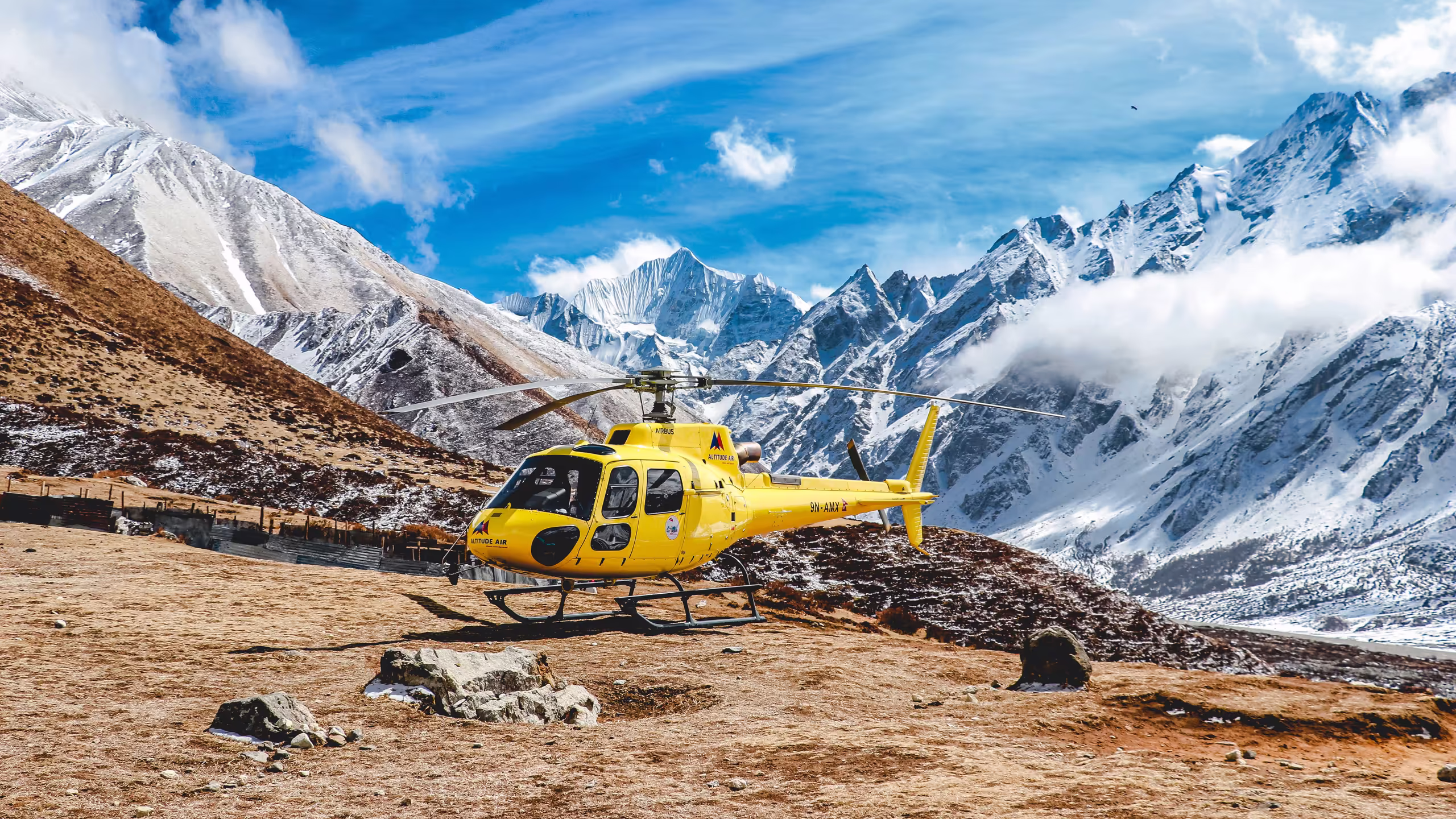 A yellow helicopter parked on a mountain landing spot with Himalayan peaks in the background
